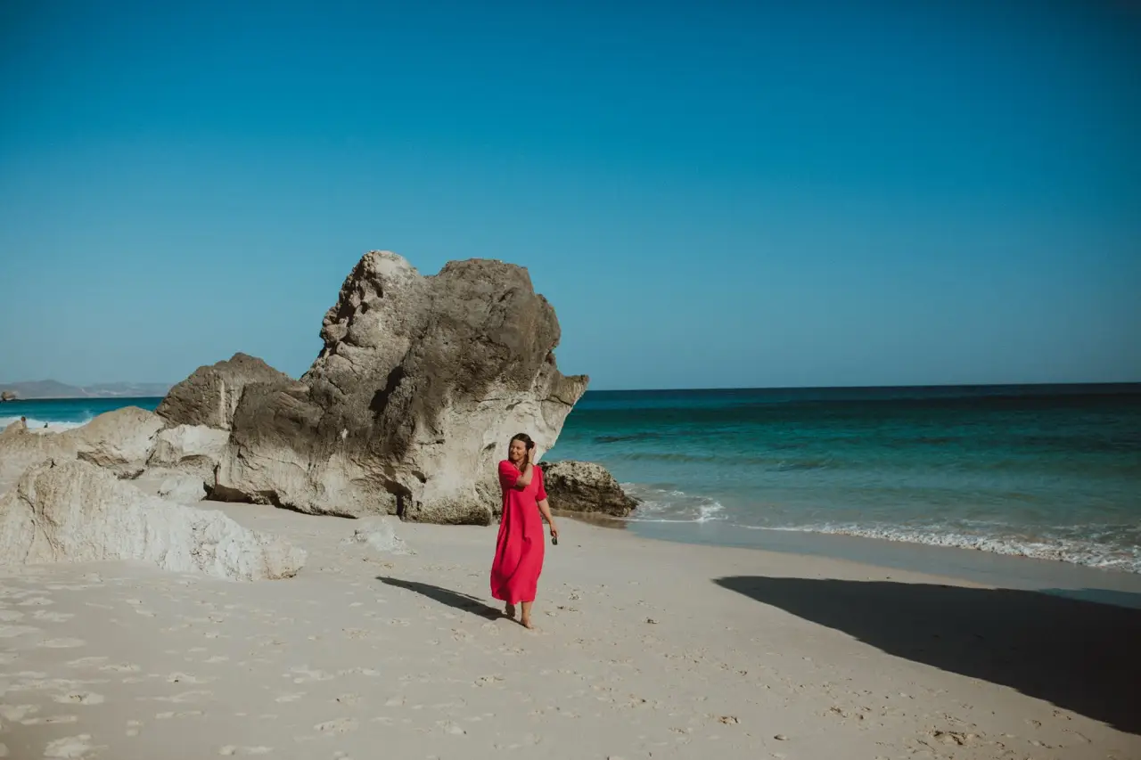 A guest in a red dress on a white-sand beach beside a large rock.