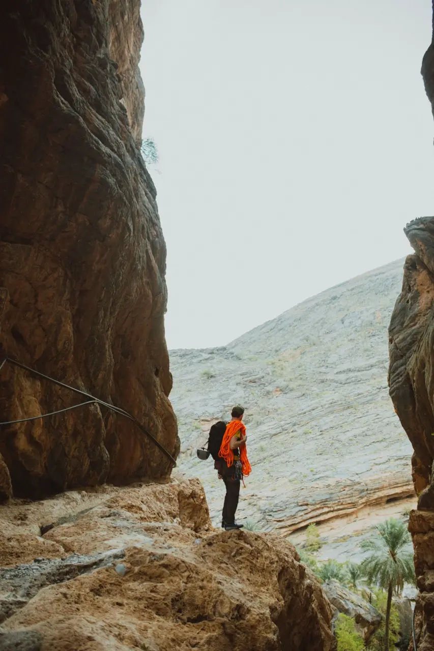 A hiker with an orange backpack on a ledge beneath a sheer canyon wall.