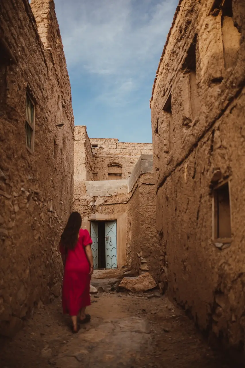 A guest in a red dress in a narrow lane between old mud-brick houses.