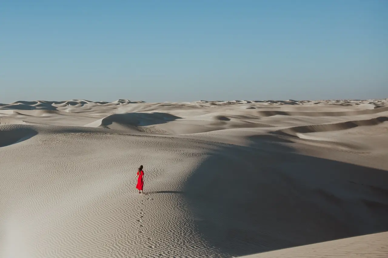 A guest in a red dress, small against wide white dunes and a blue sky.