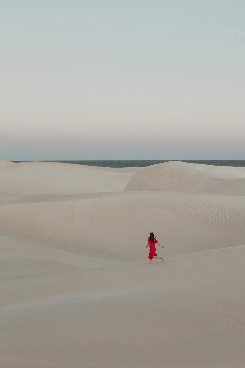 A guest in a red dress walking across pale sand dunes in the evening light.