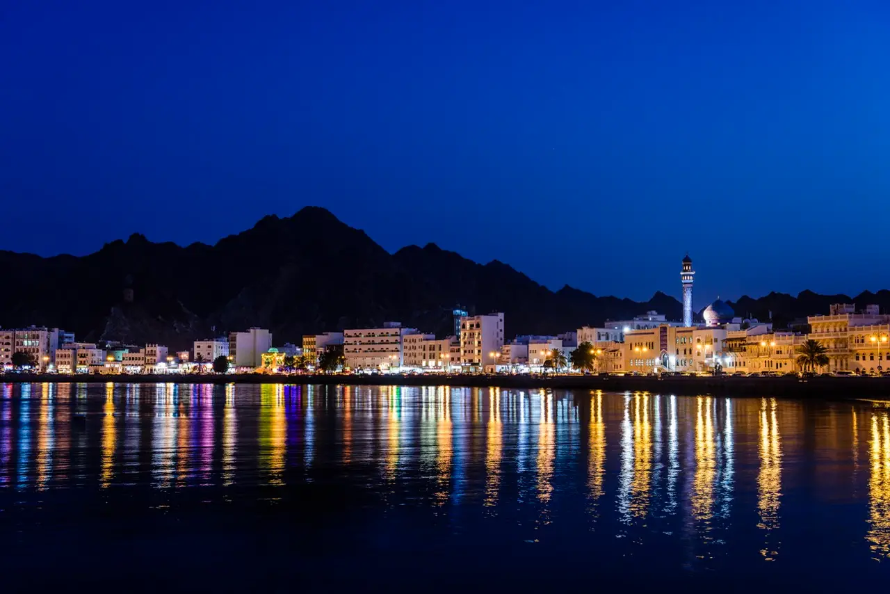 Muscat’s waterfront lit up at night below dark mountains.