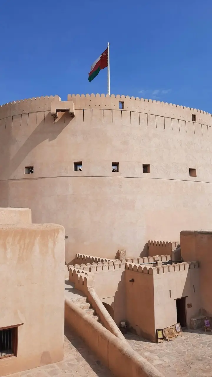 The round tower of Nizwa Fort with the Omani flag against a blue sky.